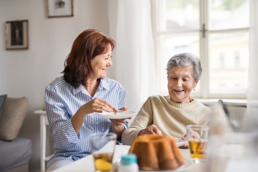 Eine Begleiterin und eine ältere Dame sitzen lächelnd beim Kaffee und Kuchen am Esstisch zusammen.